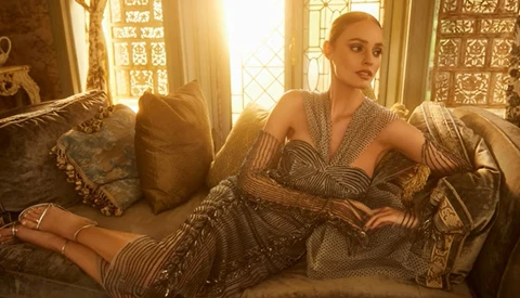 Woman in beaded wrap dress reclining on ornate sofa with decorative pillows and bright window light behind her.