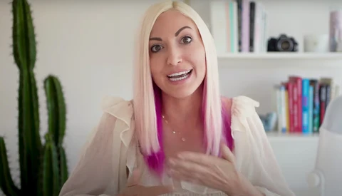 Woman with platinum blonde and pink ombre hair speaking to camera in bright, modern interior space.
