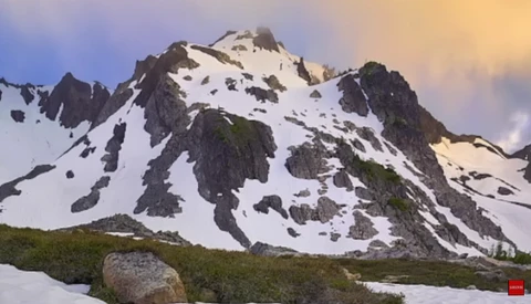 Snow-covered alpine peak with exposed dark rock faces and a lush green meadow in the foreground during golden hour.