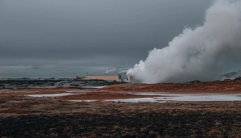 Geothermal power plant with massive white steam plumes rising from industrial structures across barren volcanic landscape.