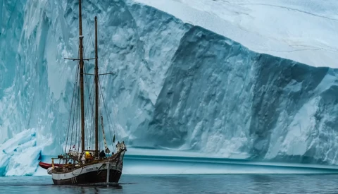 Wooden sailing vessel anchored in calm glacial waters beneath towering ice cliffs.