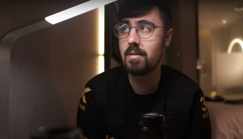 Man wearing glasses and dark clothing holds a camera in a modern interior with warm ambient lighting.
