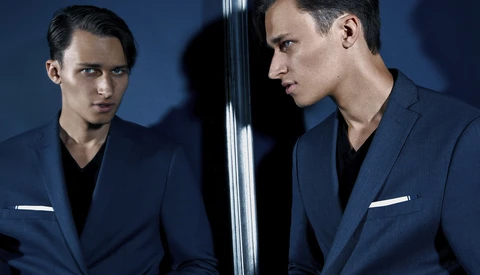 Man in navy blazer looking at his mirror reflection against blue background.