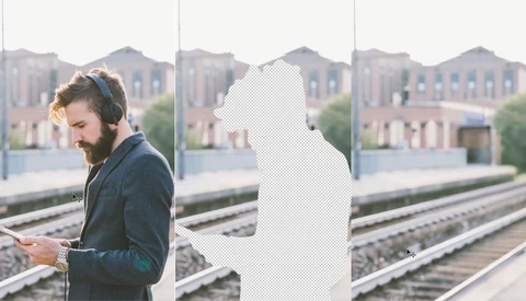 Man in blue jacket with headphones looking at phone on rooftop platform overlooking train tracks.