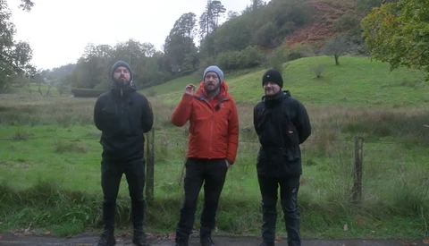 Three people in winter outdoor clothing stand in a grassy valley with forested hills in the background.