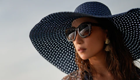 Woman wearing oversized sunglasses and a wide-brimmed navy straw hat against a bright sky.