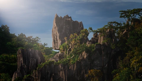 Towering limestone karst formations rise above dense tropical forest canopy beneath a partly cloudy sky.