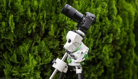 White and green motorized camera pan-tilt head mounted on tripod with DSLR camera and telephoto lens against green foliage background.