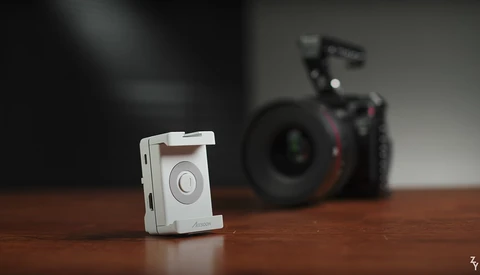 White compact camera device in sharp focus on wooden surface with professional camera equipment blurred in background.