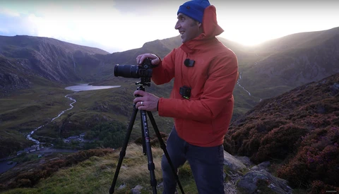 Photographer in red jacket and blue beanie operating a camera on tripod in mountainous valley landscape.