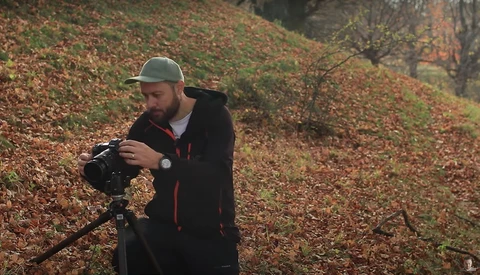 Bearded photographer adjusting a cinema camera mounted on a tripod in an autumn forest setting.