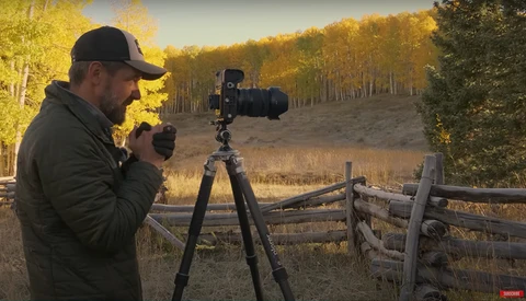 Photographer with camera on tripod composing a landscape shot during golden hour near a wooden fence.