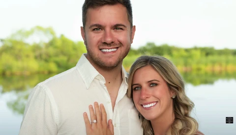 Couple posing together outdoors with water and trees in background.