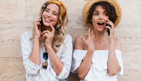 Two women wearing straw hats and white tops smiling while talking on mobile phones against a brick wall.