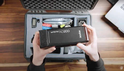Hands holding a Godox Wistro AD200 flash unit above an open equipment case on a wooden surface.
