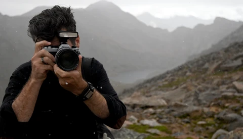 Photographer holding a DSLR camera up to his face while standing in a mountainous landscape during overcast conditions.