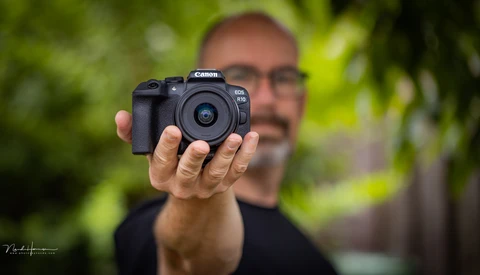 Man holding Canon EOS R50 mirrorless camera toward camera with green foliage background.