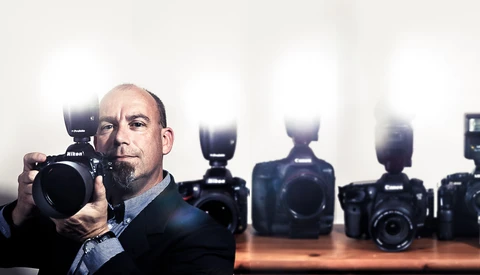 Photographer holding a Nikon DSLR camera with flash, surrounded by professional camera bodies on a wooden surface.