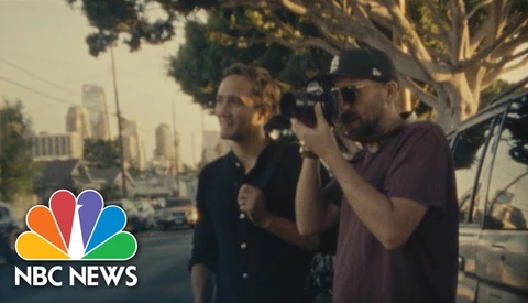 Cameraman filming two men on an urban street with city skyline in background during golden hour.