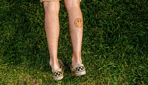 Person standing on grass wearing khaki shorts, checkered sandals, and an orange smiley face tattoo on their calf.