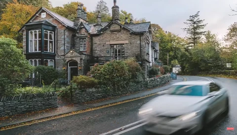 Stone mansion with bay windows beside a blurred moving car on a rural road.