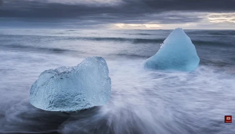 Translucent ice chunks wash ashore in shallow waves beneath a dramatic cloudy sky.