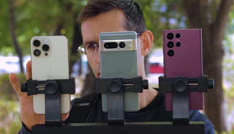 Three smartphones in different colors held by a person wearing glasses, each mounted on a black device holder for comparison.