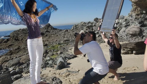 Photographer crouching to capture a portrait of a woman posing on a rocky coastal outcrop with ocean and sky in background.