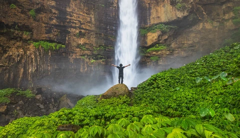 Person standing with arms raised on a rock facing a tall waterfall surrounded by lush green vegetation.