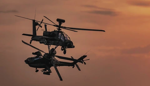Two military attack helicopters in silhouette against an orange sunset sky during flight.