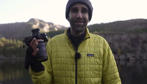 Photographer holding a telephoto lens while smiling at the camera with forested mountains and a lake visible behind him.