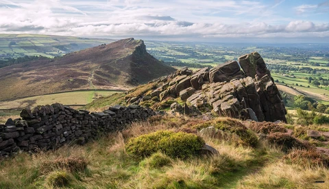 Rocky hilltop outcrop in foreground with moorland and distant peaked mountain under dramatic cloudy sky.