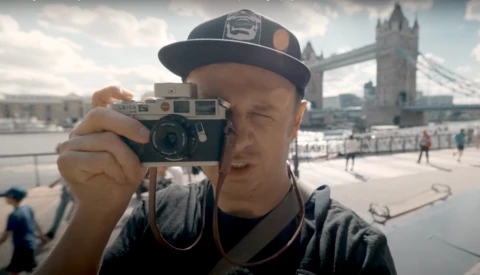 Photographer holding a vintage rangefinder camera up to his face, with Tower Bridge visible in the background.