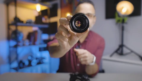 Photographer holding a vintage camera lens toward the camera in a studio environment.