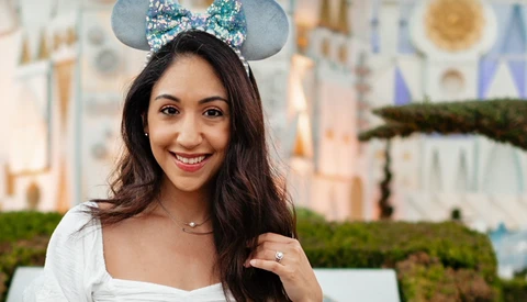 Woman wearing a sparkly blue Mickey ear headband smiles at the camera in front of a castle.