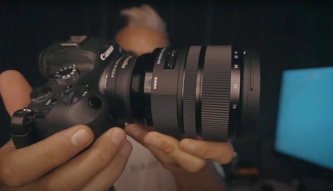 Hands holding a Canon camera with a telephoto lens attached, shown in close-up against a dark background.