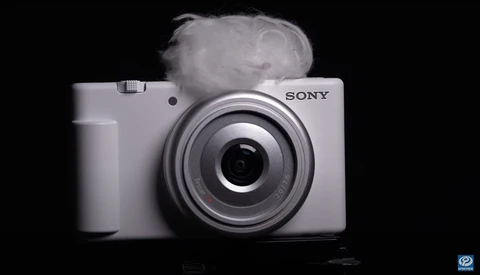 White Sony compact camera with circular lens on black background with atmospheric smoke effect.