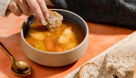 Hand dipping whole grain bread into a bowl of golden soup garnished with black pepper.