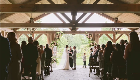 Bride and groom exchange vows under a wooden pavilion while guests watch from both sides.