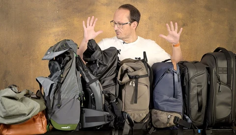 Man surrounded by camera backpacks and travel gear, hands raised in a gesture of overwhelm or indecision.