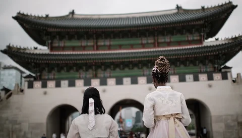 Two people in traditional white clothing viewing a large ornate Asian gateway structure with upturned eaves.