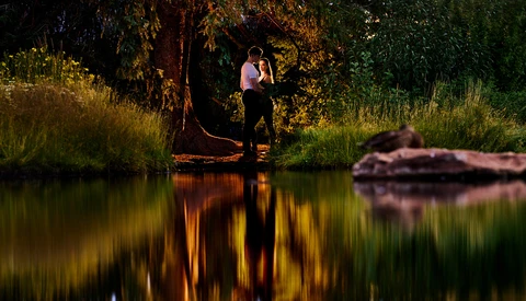 Couple embracing by a riverside at golden hour, surrounded by tall grass and trees with warm sunlight reflecting on calm water.
