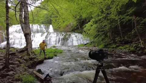 Photographer with camera on tripod capturing a multi-tiered waterfall in a lush forest stream.