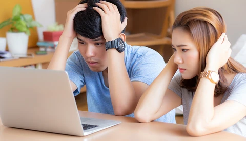 Two young adults looking at a laptop screen with concerned expressions while sitting at a wooden desk.