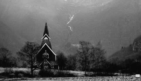 Wooden church with steeple in snowy valley beneath mountains and waterfall.