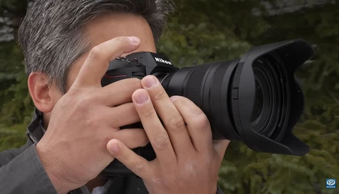 Photographer holding a Nikon DSLR camera with telephoto lens to his eye while composing a shot outdoors.