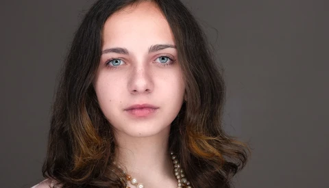 Portrait of a young woman with long dark hair and blue eyes against a neutral gray background.