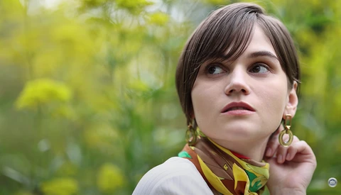 Young woman with dark hair and bangs looking upward, wearing a white shirt and patterned scarf with gold hoop earrings.