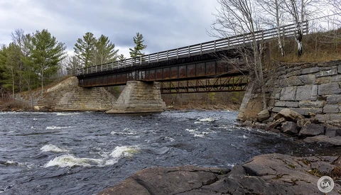 Steel truss bridge spanning a rocky river with bare trees and overcast sky in the background.