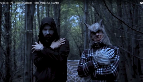 Two costumed characters in a dark forest setting, one in black robes and the other in plaid clothing with theatrical makeup.
